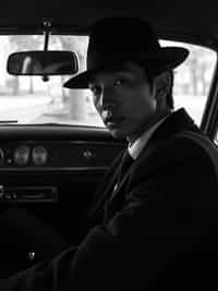 man in a vintage car interior, wearing  a dark suit with hat, dramatic lighting through car windows creating venetian blind effect, rear-view mirror showing mysterious figure, high-contrast black and white photography